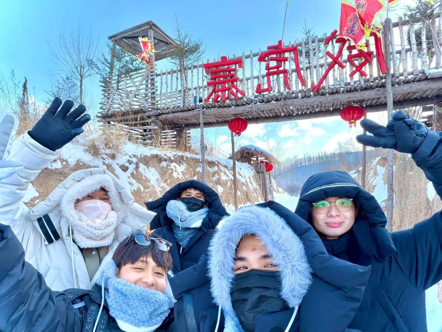The team waving in winter coats in front of a wooden gate hung with red lanterns in the snow