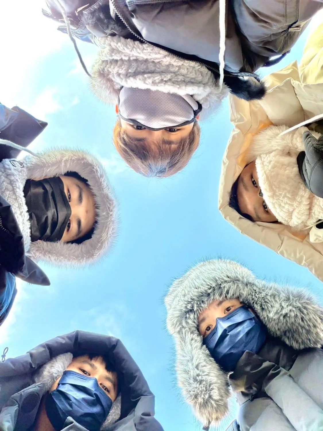 Five of the team in winter hoods, looking down into the camera in a circle against a blue sky