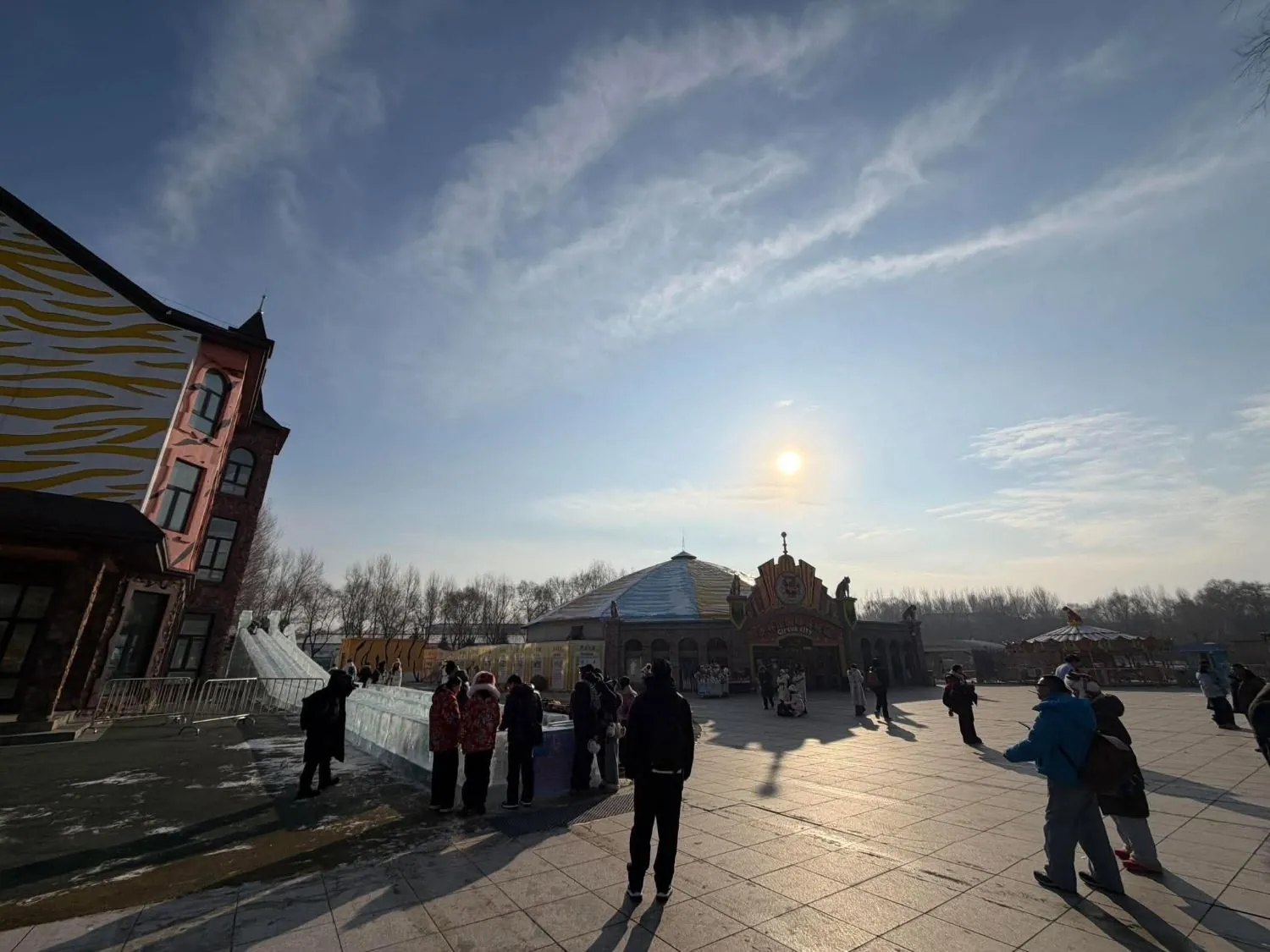 A wide square in low winter sun, with an ice slide and a round circus hall behind a crowd